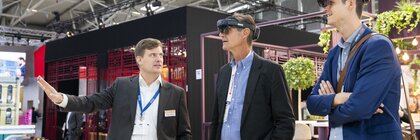 Two men in business attire stand with AR headsets at a trade fair stand and have something demonstrated by an exhibitor.