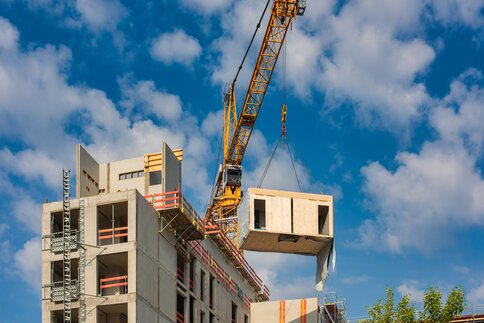 Ein Kran hebt ein Fertigteilmodul auf der Baustelle eines mehrstöckigen Gebäudes unter einem blauen Himmel mit Wolken.