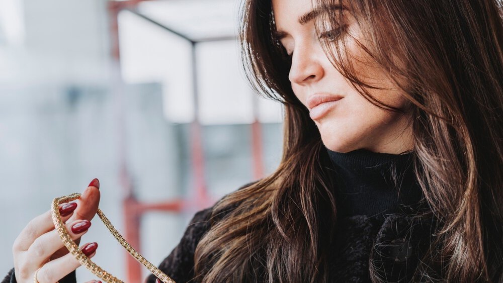 A woman with long brown hair and long red fingernails holds a gold multi-link necklace in her hands and looks at it.