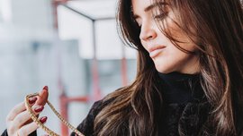 A woman with long brown hair and long red fingernails holds a gold multi-link necklace in her hands and looks at it.
