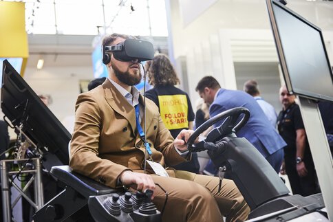 Ein Mann im braunen Anzug sitzt mit einem VR-Headset auf dem Kopf in einem Simulator, mit Lenkrad und Hebeln an einem Stand der transport logistic.