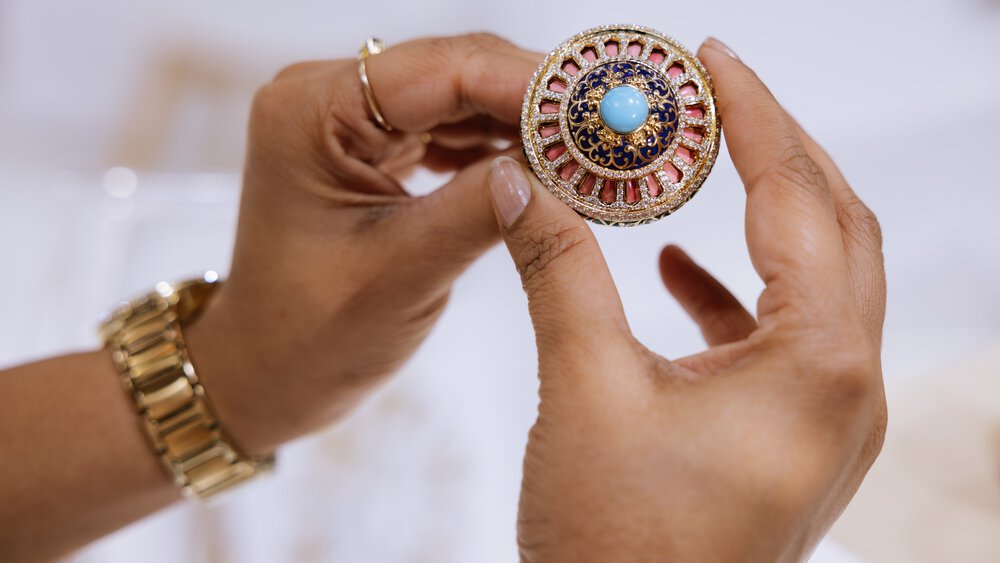  Two hands with a ring and a gold watch on the wrist hold an ornate brooch with a light blue pearl in the centre.