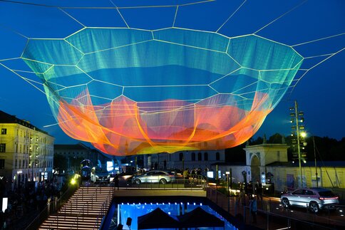 At dusk, a large, colorful net sculpture hovers over an illuminated city square, beneath which people, buildings, and cars can be seen.