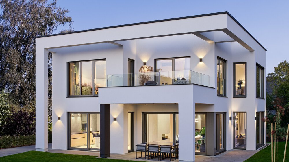 Modernes zweistöckiges Haus mit großen Fenstern, einem Balkon und Sitzplätzen im Freien, umgeben von Bäumen und einer kleinen Rasenfläche.