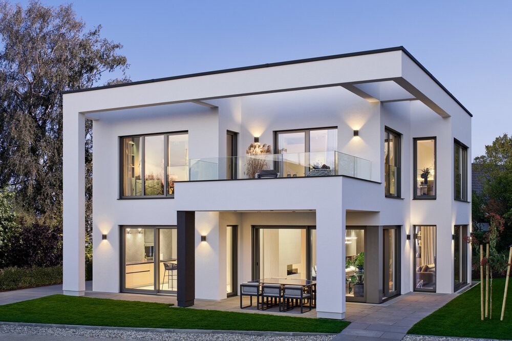 Modernes zweistöckiges Haus mit großen Fenstern, einem Balkon und Sitzplätzen im Freien, umgeben von Bäumen und einer kleinen Rasenfläche.
