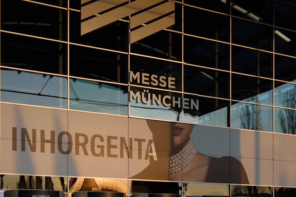 Spiegelung eines Gebäudes auf der Glasfassade der Messe München mit einer INHORGENTA-Werbung.