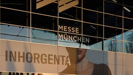 Spiegelung eines Gebäudes auf der Glasfassade der Messe München mit einer INHORGENTA-Werbung.