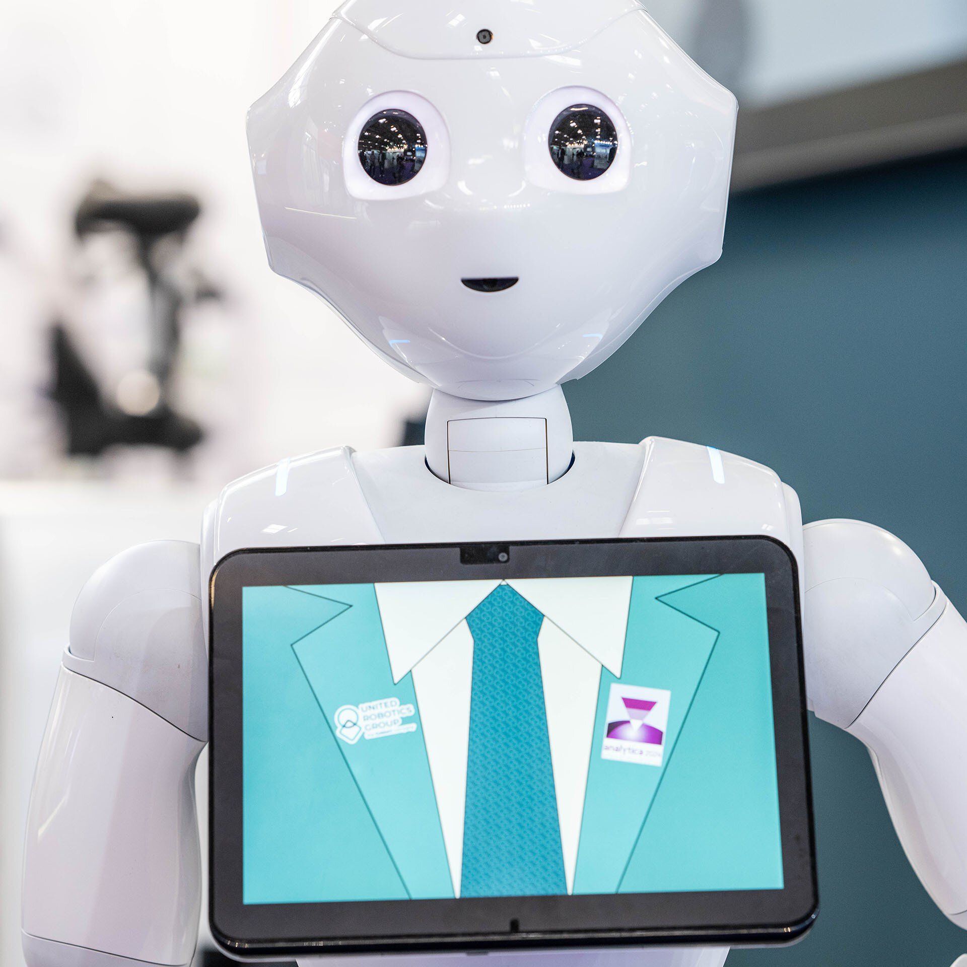 A humanoid robot with a white plastic casing has a screen on its chest. The screen shows an illustration of a suit and tie and the analytica logo.