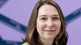 Eine lächelnde Frau mit schulterlangen braunen Haaren, einer weißen Bluse und einem hellgrünen Blazer steht vor einem lila Hintergrund.