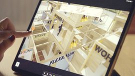 Nahaufnahme eines eingeschalteten Tablets, auf dessen Bildschirm ein Modell aus mehreren gestapelten Holzräumen mit teils offenen Wänden zu sehen ist.