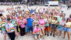 Eine große Gruppe von lächelnden Menschen mit Regenbogenfächern bei einer Veranstaltung im Freien.