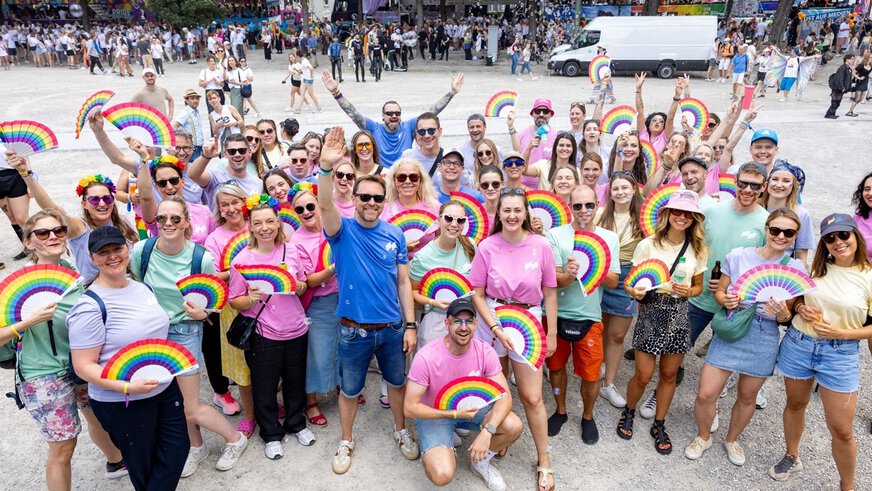 Eine große Gruppe von lächelnden Menschen mit Regenbogenfächern bei einer Veranstaltung im Freien.