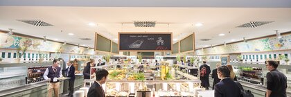 Trade fair visitors with tablets walk through a brightly lit self-service counter with drinks and food of all kinds.