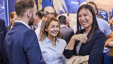 Zwei Frauen in Businesskleidung unterbrechen ihre Unterhaltung mit einem Mann, um für das Foto zu lächeln. Im Hintergrund sind weitere Besucher.