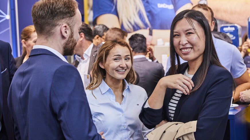 Zwei Frauen in Businesskleidung unterbrechen ihre Unterhaltung mit einem Mann, um für das Foto zu lächeln. Im Hintergrund sind weitere Besucher.