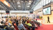 Many people sit in an exhibition hall facing a stage and listening to a presentation, with a speaker standing to the right next to a large screen.         