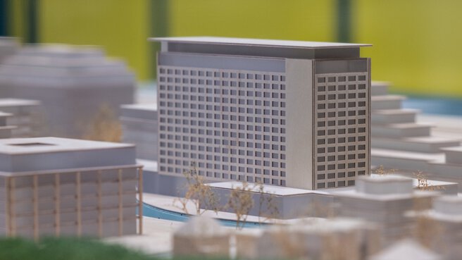 Close-up of an architectural model with focus on a modern high-rise surrounded by smaller buildings and trees.