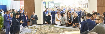 Besucherinnen und Besucher der EXPO REAL in München betrachten ein großes, beleuchtetes Stadtmodell an einem Messestand. Mehrere Personen diskutieren über das Modell, einige machen Fotos oder hören Präsentationen zu. Im Hintergrund sind viele weitere Messegäste, Gesprächsgruppen und Ausstellerstände zu sehen, die eine lebhafte Atmosphäre schaffen.