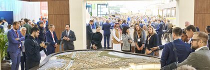 Besucherinnen und Besucher der EXPO REAL in München betrachten ein großes, beleuchtetes Stadtmodell an einem Messestand. Mehrere Personen diskutieren über das Modell, einige machen Fotos oder hören Präsentationen zu. Im Hintergrund sind viele weitere Messegäste, Gesprächsgruppen und Ausstellerstände zu sehen, die eine lebhafte Atmosphäre schaffen.