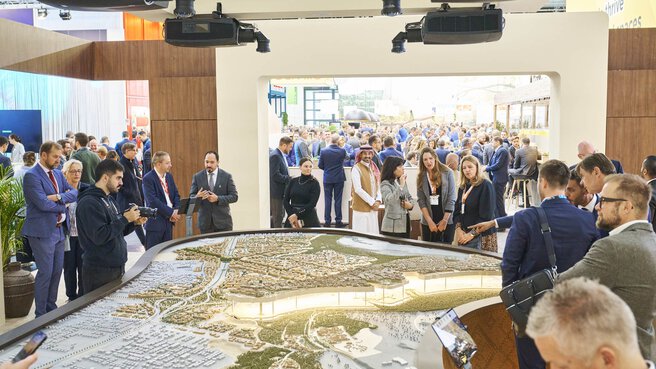 Besucherinnen und Besucher der EXPO REAL in München betrachten ein großes, beleuchtetes Stadtmodell an einem Messestand. Mehrere Personen diskutieren über das Modell, einige machen Fotos oder hören Präsentationen zu. Im Hintergrund sind viele weitere Messegäste, Gesprächsgruppen und Ausstellerstände zu sehen, die eine lebhafte Atmosphäre schaffen.