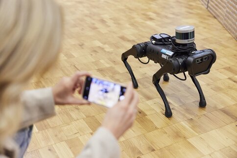Someone takes a photo of a small, black robot dog with a sensor, on a wooden floor.
