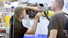 Zwei Männer arbeiten gemeinsam an einem Industrieroboter auf einer Messe. Einer hält Werkzeuge und justiert eine mechanische Komponente, während der andere beobachtet.