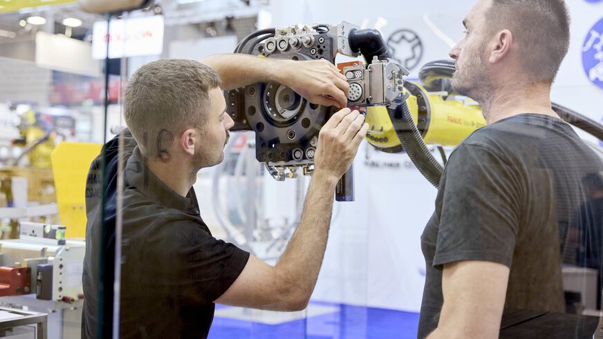 Zwei Männer arbeiten gemeinsam an einem Industrieroboter auf einer Messe. Einer hält Werkzeuge und justiert eine mechanische Komponente, während der andere beobachtet.