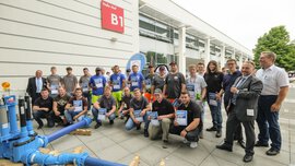 A group of people in work clothes posing with awards in their hands next to a blue pipe installation in front of exhibition hall B1.