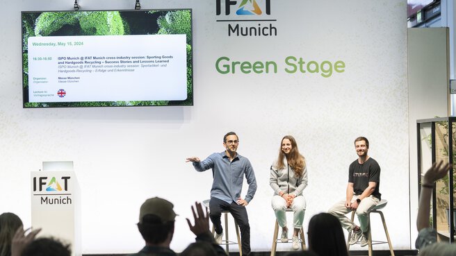 Three people sitting on the IFAT Munich Green Stage presenting,  while the members in the audience raise their hands.