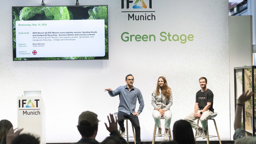Three people sitting on the IFAT Munich Green Stage presenting,  while the members in the audience raise their hands.