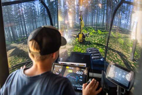 Junge steuert einen Forstmaschinen-Simulator und sieht auf einem großen Bildschirm eine virtuelle Waldszene.