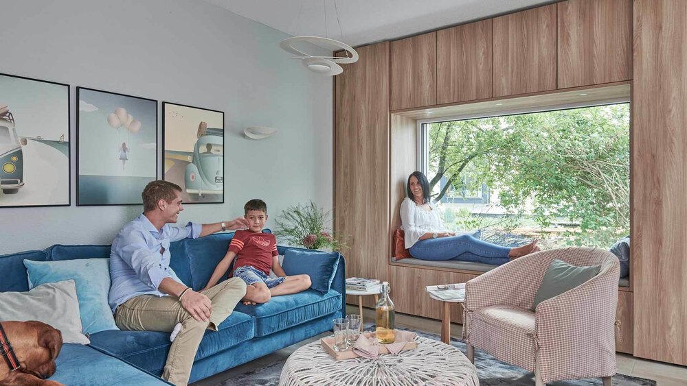 Eine Familie in einem modernen Wohnzimmer mit großem Fenster, blauem Sofa und Hund. Eine Frau sitzt auf dem Fensterplatz.