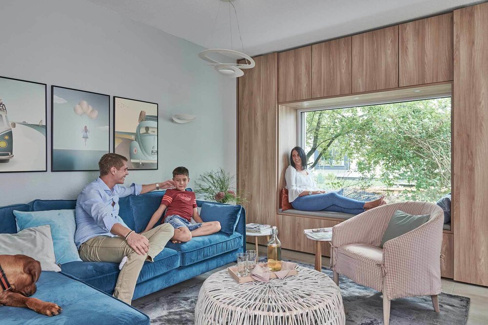 Eine Familie in einem modernen Wohnzimmer mit großem Fenster, blauem Sofa und Hund. Eine Frau sitzt auf dem Fensterplatz.