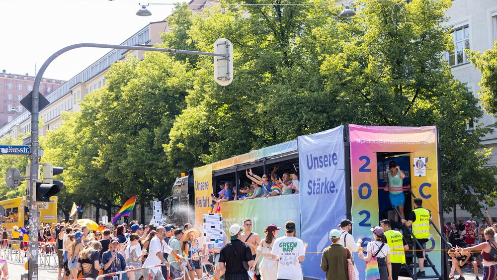 A colorful parade float bearing the slogan “Our Strength” passes by spectators at a sunny open-air festival or a Pride event.