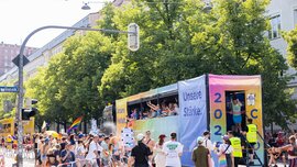 A colorful parade float bearing the slogan “Our Strength” passes by spectators at a sunny open-air festival or a Pride event.