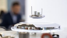 Nahaufnahme einer runden liegenden Lampe, darüber schwebt ein kleines technisches Gerät. Im Hintergrund sieht man verschwommen eine Person.