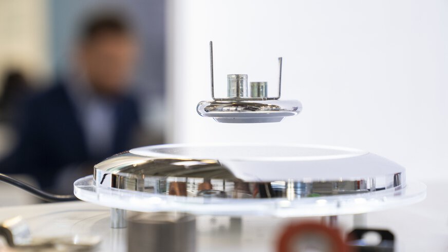 Nahaufnahme einer runden liegenden Lampe, darüber schwebt ein kleines technisches Gerät. Im Hintergrund sieht man verschwommen eine Person.