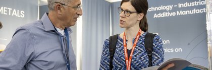 Eine Frau stellt einem Aussteller an einem Messestasnd Fragen zu einer Broschüre, die sie in den Händen hält.
