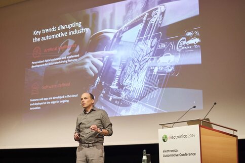 A man speaks on the electronica stage. He is standing next to the podium and a slide about the automotive industry can be seen on the screen behind him.