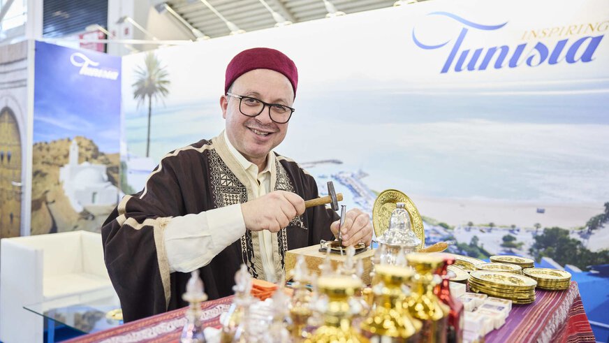 Traditionally dressed man at a Tunisia stand at a travel fair, artfully working sheet metal.