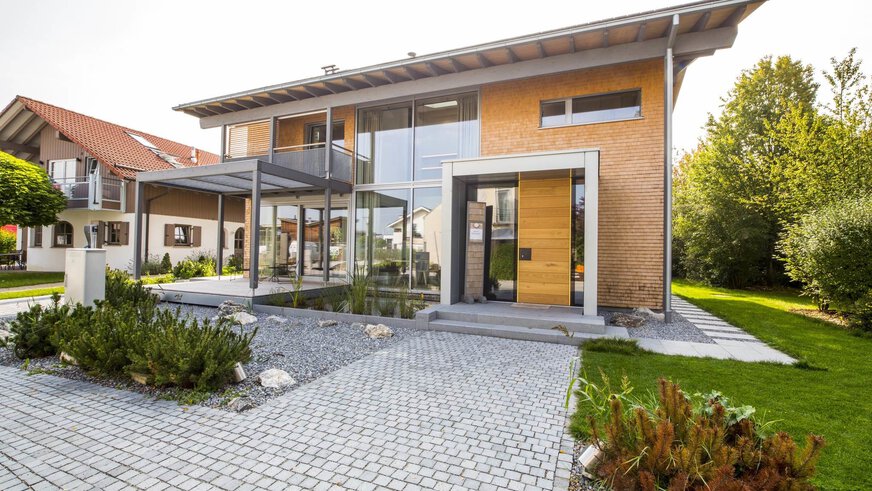 Modernes zweistöckiges Haus mit großen Fenstern, umgeben von Grün und einem gepflegten Garten.