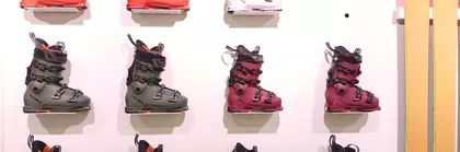 Elf Skischuhe in verschiedenen Farben und zwei gelbe Skier werden an einer Ausstellerwand präsentiert.