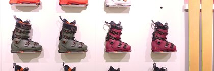 Elf Skischuhe in verschiedenen Farben und zwei gelbe Skier werden an einer Ausstellerwand präsentiert.