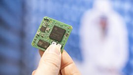 Nahaufnahme einer Hand, die eine grüne Leiterplatte mit sichtbaren Mikrochips vor einem unscharfen blauen Hintergrund hält.
