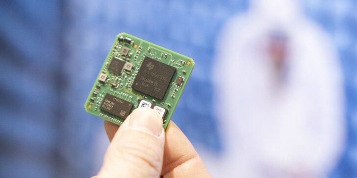Close-up of a hand holding a green circuit board with visible microchips in front of a blurred blue background.