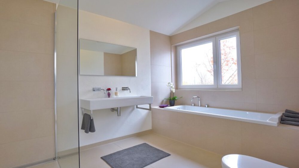 Minimalistisches Badezimmer mit wandmontiertem Waschbecken, Badewanne, großem Spiegel und Fenster. Beige Wände und ein einfacher grauer Teppich.