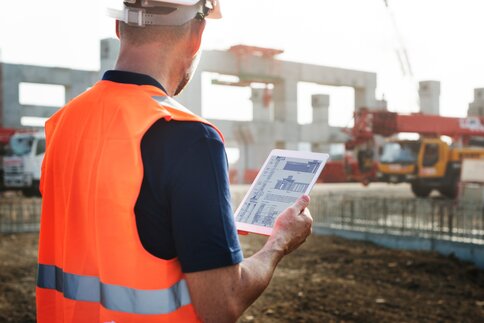 Bauarbeiter im orangenen Schutzanzug und Helm betrachtet Baupläne auf einem Tablet auf einer Baustelle.
