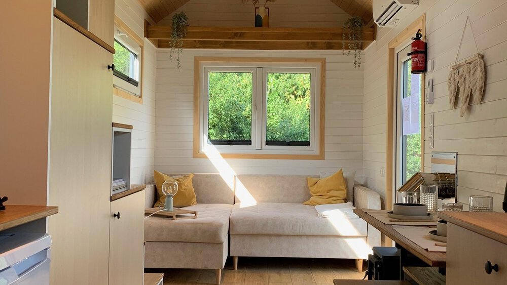 Gemütlicher Wohnbereich im Tiny House mit beigem Sofa, großem Fenster und Holzbalken an der Decke.