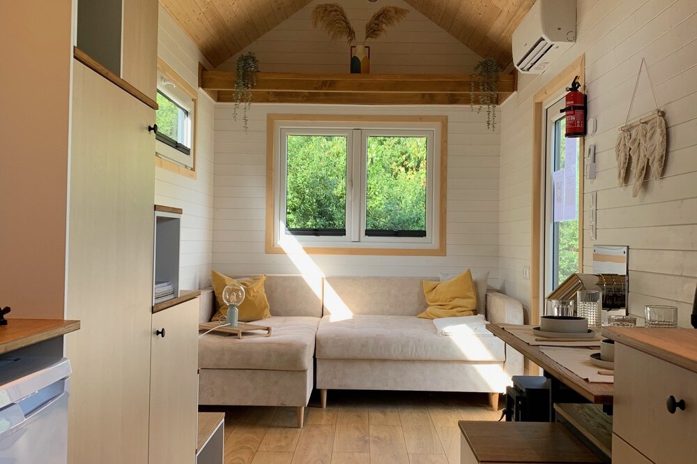 Gemütlicher Wohnbereich im Tiny House mit beigem Sofa, großem Fenster und Holzbalken an der Decke.