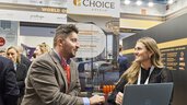 A woman and a man are smiling and talking at a Choice Hotels trade fair booth. A laptop sits in front of them, with other attendees and company promotional displays visible in the background.